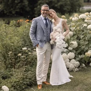 Un couple de mariés pose avec élégance dans un jardin fleuri. Le marié porte un smoking moderne et raffiné de la boutique Surmesur, catégorie Smoking de mariage, avec une veste à carreaux bleu-gris, un gilet assorti et un pantalon beige. Il est complété par des chaussures marron élégantes. La mariée est vêtue d