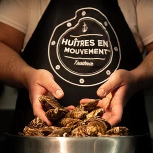 Une image montrant un traiteur nommé Oysters In Motion, spécialisé dans les mariages, exposant fièrement des huîtres fraîches dans ses mains. Le chef porte un tablier noir avec le logo de l