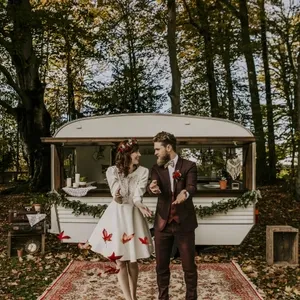 Un couple de mariés se tient devant une caravane décorée, entourée de forêt. La mariée porte une robe courte avec des manches longues en dentelle et une couronne de fleurs rouges. Le marié, habillé par Blandin & Delloye, porte un élégant smoking de mariage bordeaux assorti à sa boutonnière. L