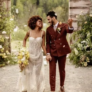 Un couple élégant sourit en se tenant la main, vêtu pour un mariage. La mariée porte une robe pailletée sans manches avec un bouquet de fleurs jaunes, tandis que le marié, Blandin & Delloye, porte un smoking de mariage bordeaux avec une boutonnière florale. Ils sont debout devant une arche en pierre, entourée de verdure et de fleurs, créant une ambiance romantique et naturelle. Cette image illustre parfaitement un moment de joie et de complicité pendant une cérémonie de mariage.