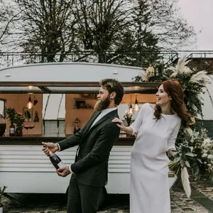 Un couple joyeux devant une caravane décorée pour un événement de mariage célèbre le grand jour. Le marié, vêtu d