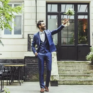 Un homme élégant porte un smoking bleu de mariage, conçu par Blandin & Delloye, dans la catégorie Smoking de mariage. Il se tient dans une cour extérieure bordée de verdure. Son allure est sophistiquée, avec une chemise blanche et une cravate assortie. L