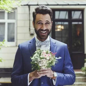 Un homme souriant portant un élégant smoking bleu, fourni par Blandin & Delloye, se tient devant un bâtiment charmant. Il porte une chemise blanche et un nœud papillon assorti, ce qui ajoute une touche sophistiquée à l