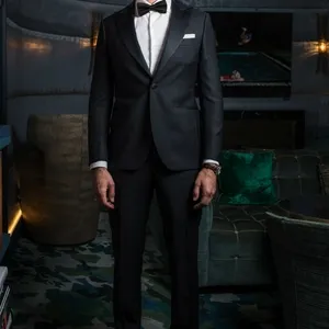 Un homme élégant porte un smoking de mariage noir, confectionné par Blandin & Delloye, catégorie smoking de mariage. Le smoking comprend une veste parfaitement ajustée, une chemise blanche impeccable et un nœud papillon noir. Il se tient debout dans un salon raffiné, avec des fauteuils en velours et un tapis sophistiqué au motif unique, ajoutant une ambiance luxueuse, idéale pour une réception de mariage ou un événement haut de gamme.