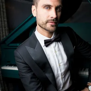 Homme élégant portant un smoking de mariage noir avec un nœud papillon, fourni par Blandin & Delloye, assis devant un piano à queue. Parfait pour les mariages sophistiqués, ce smoking incarne le style et le raffinement.