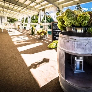 La Distinction, salle de réception, présente un élégant espace en plein air avec une terrasse couverte en bois offrant une vue panoramique sur la nature luxuriante environnante. Des tables hautes et des chaises blanches sont disposées harmonieusement pour accueillir les invités. Au premier plan, un tonneau en bois sert de décoration rustique, et des arrangements floraux de couleurs vives ajoutent une touche festive. Une ardoise détaillant le menu ou le programme de l