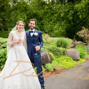 Un couple de mariés souriants se tient dans un jardin verdoyant, tenant chacun un verre de cocktail. La mariée porte une robe blanche ornée de dentelle et un voile, tandis que le marié est vêtu d