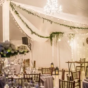La salle de réception La Distinction présente un espace élégant avec de longues tables drapées de tissus blancs et décorées de bouquets luxuriants de fleurs blanches. Des chaises en bois sombre entourent les tables, et des verres à pied sont soigneusement disposés pour le service. Un grand lustre scintillant illumine l