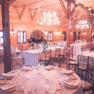 La salle de réception La Distinction présente un décor enchanteur avec un plafond en bois, des lustres élégants et des guirlandes lumineuses créant une ambiance chaleureuse. Les tables sont magnifiquement dressées avec des nappes blanches, des verres à pied et des couverts soigneusement disposés. De superbes compositions florales ornent chaque table, ajoutant une touche de sophistication à cet espace destiné à accueillir des mariages romantiques.