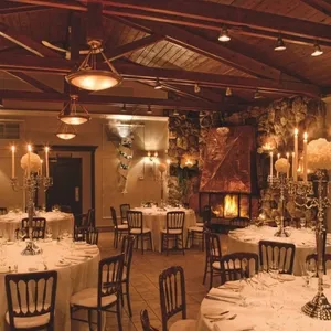 La salle de réception La Distinction présente une atmosphère élégante et accueillante, idéale pour un mariage. La pièce est décorée avec des tables rondes, chacune drapée de nappes blanches impeccables, et ornée de chandeliers en argent sophistiqués. Des bougies surélevées ajoutent une touche de charme romantique. Le plafond en bois avec des poutres apparentes amplifie le caractère rustique-chic de l