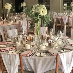 La Distinction, une salle de réception élégante, est présentée avec un décor de mariage raffiné. La scène montre des tables magnifiquement dressées avec des nappes blanches, des serviettes roses délicatement pliées, et de chaises en bois recouvertes de housses blanches transparentes. Chaque table est ornée d
