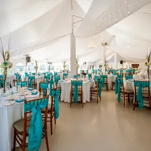 Dans cette image, la salle de réception La Distinction est élégamment décorée pour un mariage. Les tables rondes sont drapées de nappes blanches impeccables, accentuées par des chemins de table turquoise éclatants. Chaque chaise est ornée d