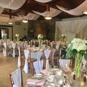 La Distinction, salle de réception, présente une élégante salle ornée de drapés blancs suspendus au plafond, créant une ambiance romantique. Les tables rondes sont magnifiquement décorées avec des nappes blanches et des serviettes roses, agrémentées de hauts bouquets de fleurs blanches et vertes comme centre de table. Les chaises en bois sont habillées de housses blanches et de nœuds élégants. La lumière douce et les guirlandes lumineuses ajoutent à l