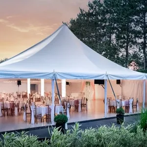 Sous un ciel de coucher de soleil, une élégante tente blanche est installée dans un cadre extérieur serein pour servir de salle de réception. Cette structure appartient à La Distinction, un prestataire spécialisé dans les salles de réception pour mariages. La tente est aménagée avec des tables décorées de nappes blanches et entourées de chaises en bois, prêtant une atmosphère raffinée et accueillante. De la verdure environne l