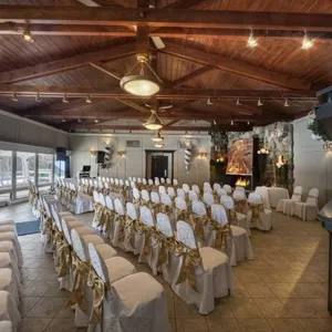 La salle de réception de La Distinction, spécialisée dans les mariages, présente une élégante configuration avec des chaises alignées et recouvertes de housses blanches et de nœuds dorés. La pièce est dotée d