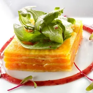 Dans cette image de La Distinction, catégorie Salle de réception, on voit un plat élégant présenté sur une assiette blanche. Au centre, une structure carrée composée de fines tranches de légumes jaunes est garnie de feuilles de verdure fraîches. Autour du carré, une sauce rouge est délicatement disposée en cercle, ajoutant une touche de couleur et de sophistication. Ce type de présentation culinaire raffinée est idéal pour un cadre de mariage élégant, mettant en valeur l