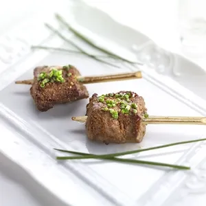 Deux délicieuses brochettes de viande décorées de ciboulette fraîche sont soigneusement présentées sur une assiette blanche élégante, avec des brins de ciboulette disposés artistiquement. Cette image illustre le raffinement culinaire offert par La Distinction, une salle de réception spécialisée en organisation de mariages, inscrite sur Mariage Elegant, Canada.