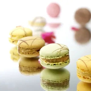 Macarons délicats disposés sur une surface réfléchissante, capturant les nuances pastel du jaune, vert, et rose. Ces gourmandises élégantes sont parfaites pour embellir un événement chez La Distinction, une salle de réception renommée. En arrière-plan, de légères touches de couleur ajoutent une ambiance festive, renforçant le cadre idéal pour un mariage raffiné.